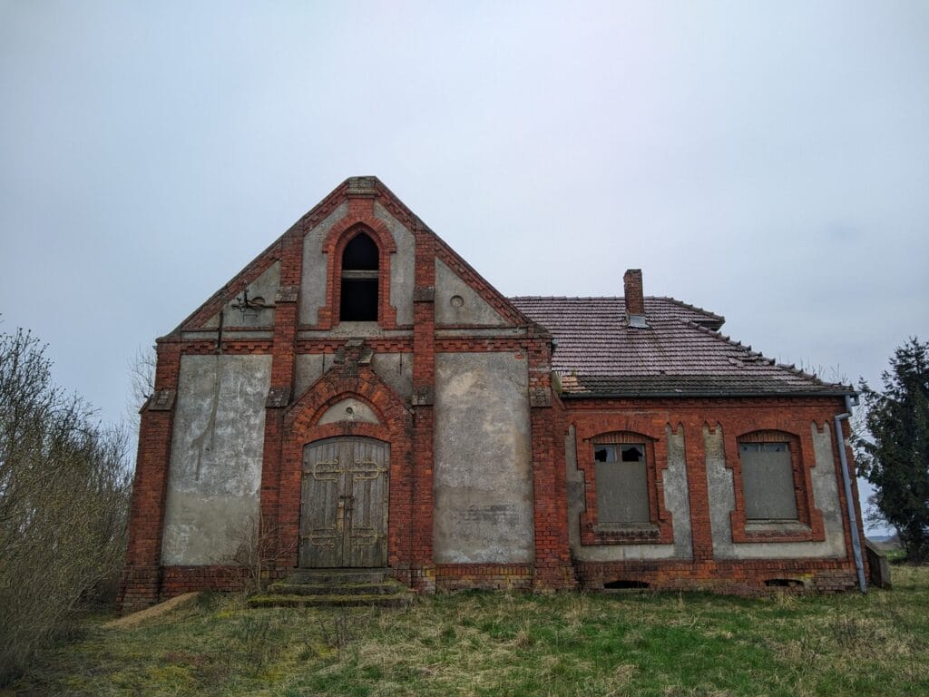 Budynek kaplicy widziany od frontu. Po lewej główne wejście do sali modlitewnej, po prawej niższe skrzydło pastorówki. Budynek z czerwonej cegły, otynkowany między wystającymi elementami (lizenami).
