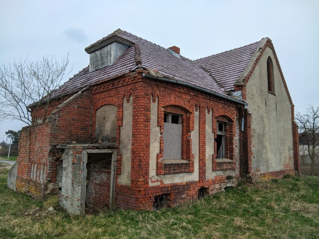 Tył budynki, widoczne mocno zniszczone zejście do piwnicy znajdujące się pod schodami do pastorówki. Okna pastorówki bez szyb. Jedno z nich częściowo załonięte płytą.