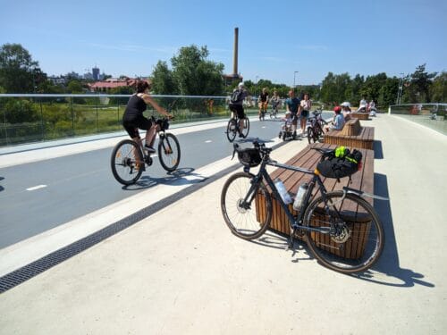 Mój rower oparty o drewnianą ławkę stojącą na pieszej części mostu dla pieszych i rowerów. Na sąsiedniej ławce siedzą ludzie w kaskach rowerowych. Po prawej kilka osób jedzie na rowerach, w środku idzie rodzina z dzieckiem na małym trójkołowym rowerku, dalej po prawej dwie osoby z wózkami i dziecięcymi.