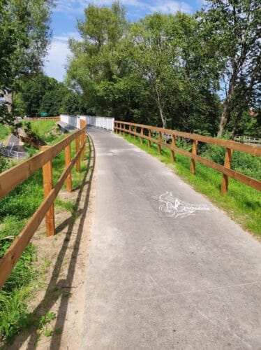 Otoczony drewnianymi barierkami podjazd na krótki most. Po bokach asfaltowej drogi dla rowerów trawa i drzewa.