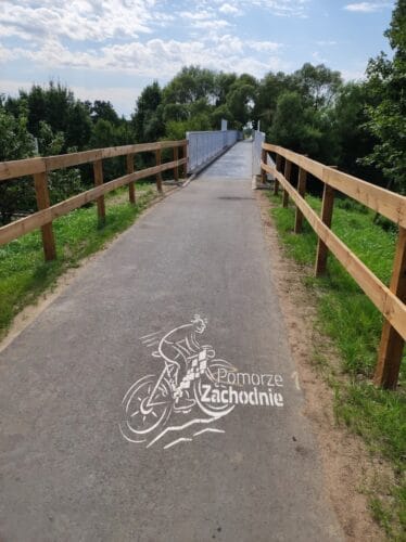 Wjazd na drugi, dłuższy most dla rowerów. Również z drewnianymi barierkami. Na asfalcie namalowany białą farbą pędzący rowerzysta i napis Pomorze Zachodnie.