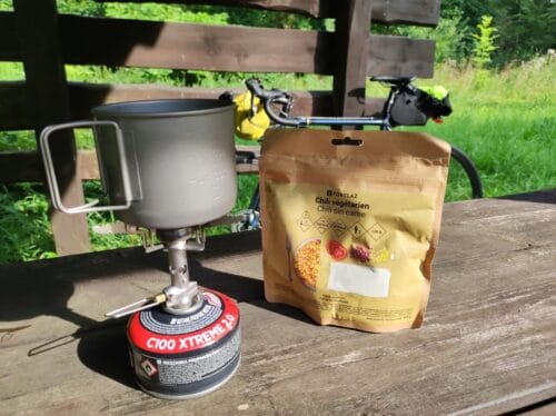 Na drewnianym stole pod wiatą stoi nieduża kuchenka gazowa, na której gotuje się woda w metalowym kubku. Obok paczka liofilizowanego wegetariańskiego chilli. W tle wystaje kawałek ciemnoniebieskiego roweru.