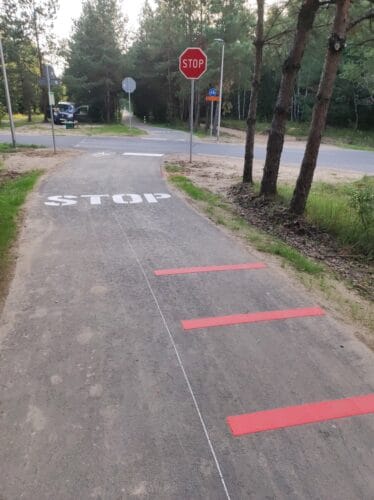 Droga dla rowerów dochodzącą do skrzyżowania z szosą. Na DDR nałożone są czerwone wypukłe paski w poprzek i duży biały napis stop. Obok tego czerwony znak drogowy stop.