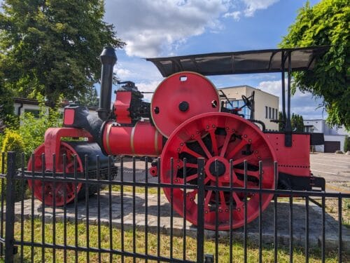 Stary, czerwono-czarny walec parowy stoi za czarnym metalowym płotem.
