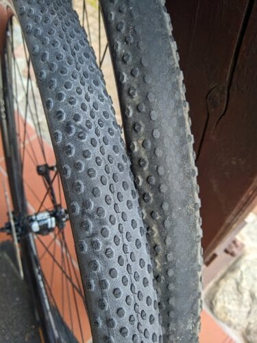 Zbliżenie na opony na dwóch kołach rowerowych opartych o siebie. Lewa opona jest nowa, z wyraźnym bieżnikiem, ta po prawej ma klocki na środku wytarte na gładko.