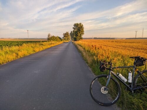 Szosa między polami oświetlonymi złotym światłem porannego słońca. Po prawej stronie stoi ciemnoniebieski rower szutrowy, oparty o znajdujący się poza kadrem słupek.