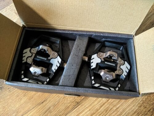 W kartonowym pudełku, którego wierzchnia strona jest pomalowana na ciemnoszary z ciemniejszymi kropkami i kreskami, leżą dwa pedały.