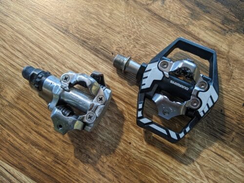 Dwa pedały leżą na blacie stołu. Po lewej wyraźnie używane pedał, minimalny, z samą częścią do wpinania bloków znajdujących się na spodzie rowerowych butów. Po lewej nowiutkie, z czarną metalową klatką dookoła.