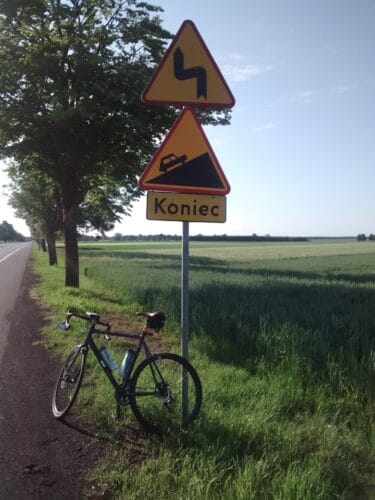 Na trawiastym poboczu przy szosie stoi znak oznaczający koniec ciągu niebezpiecznych zakrętów i stromego podjazdu. O znak oparty jest tylnym kołem ciemnoniebieski rower gravelowy.