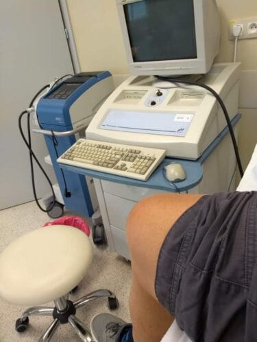 Noga w krótkich spodenkach na tle jakiegoś medycznego urządzenia z monitorem, klawiaturą i myszką.