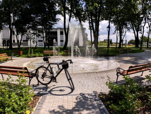 Alejka z prowadzi do niedużej fontanny w parku. Po jej bokach stoją ławki. O ławkę po lewej oparty jest tylnym kołem ciemnoniebieski rower szutrowy.
