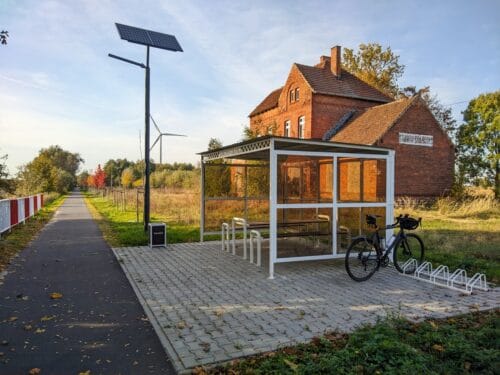 Przy równej, asfaltowej drodze dla rowerów stoi przeszklona wiata ze stołem i ławami. W stojaku obok niej stoi rower szutrowy.