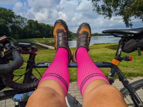 Nogi w jaskrawych różowych podkolanówkach i pomarańczowo-czarnych butach opierają się o ramę ciemnoniebieskiego roweru. W tle widać jezioro.