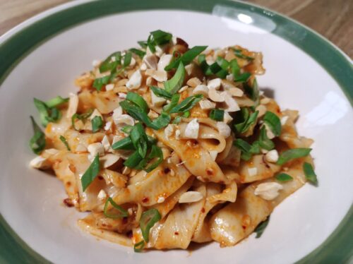 W białej misce z szerokim zielonym paskiem dookoła brzegu znajduje się porcja szerokiego makarony w delikatnym czerwono-pomarańczowym sosie z widocznymi płatkami chilli. Na wierzchu cięty po skosie szczypior i nierówno posiekane orzeszki ziemne.