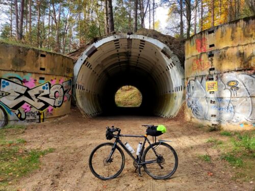 Na tle betonowej konstrukcji sporego, ale krótkiego, okrągłego tunelu stoi ciemnoniebieski rower szutrowy, pobrudzony błotem. Ściany po bokach tunelu pomazane kolorowym graffiti.
