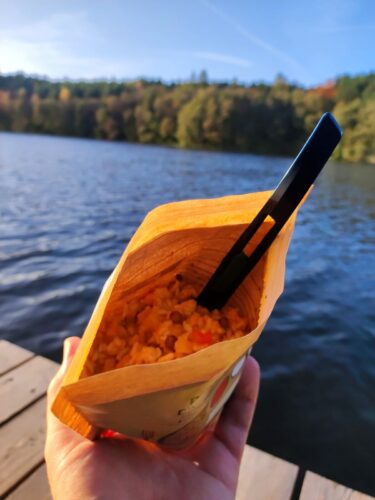 Trzymana w ręce paczka z gotowym daniem, w tle kawałek pomostu, tafla jeziora i ściana lasu.