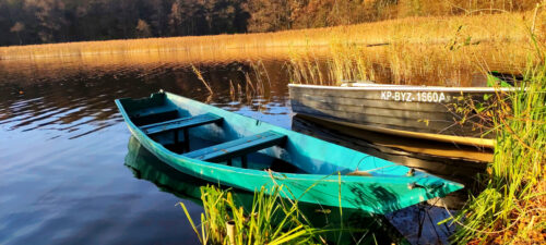Kolejne dwie łodzie przycumowane przy brzegu jeziora. Bliższa jest drewniana i jaskrawo zielona, dalsza czarna i z tworzywa.