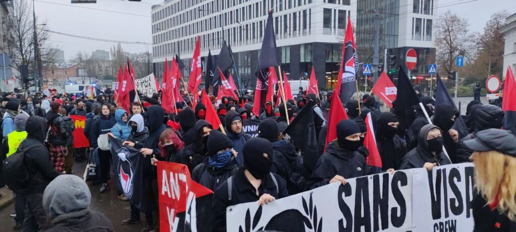 Demonstracja. Większą część zdjęcia zajmuje grupa ubranych na czarno osób w kominarkach z czerwonymi i czarnymi flagami. Osoby z przodu trzymają transparent z napisem „Sans visage”