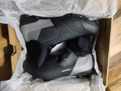 W kartonie wysłanym białym pergaminem leżą dwa czarne buty za kostkę z odblaskowymi piętami. Napisy na nich to: „BOA”, „Shimano” oraz „MW7”.