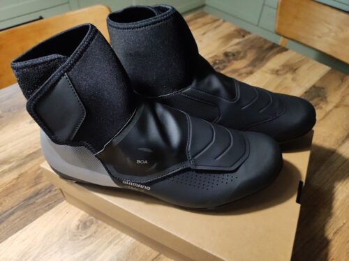 Te same dwa buty teraz stoją na zamkniętym kartonie leżącym na drewnianym stole.