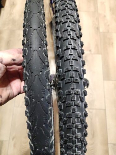 Dwie opony rowerowe obok siebie. Lewa jest trochę węższa, ma kostki bieżnika tylko po brzegach, a środek jest prawie całkiem gładki, z lekkimi rowkami. Opona po prawej jest szersza i ma duże kostki bieżnika na całej szerokości.