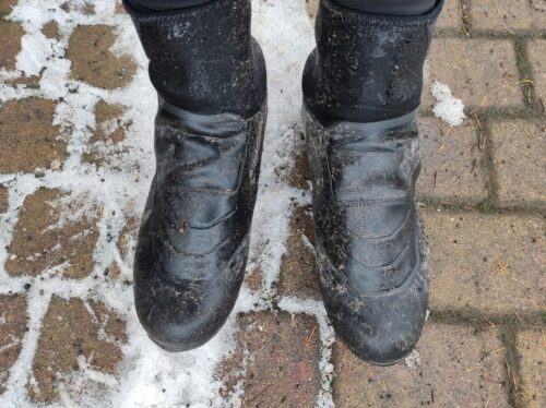 Na kostce brukowej z resztkami śniegu stoją dwie nogi w butach zimowych Shimano, mokrych i umazanych błotem.