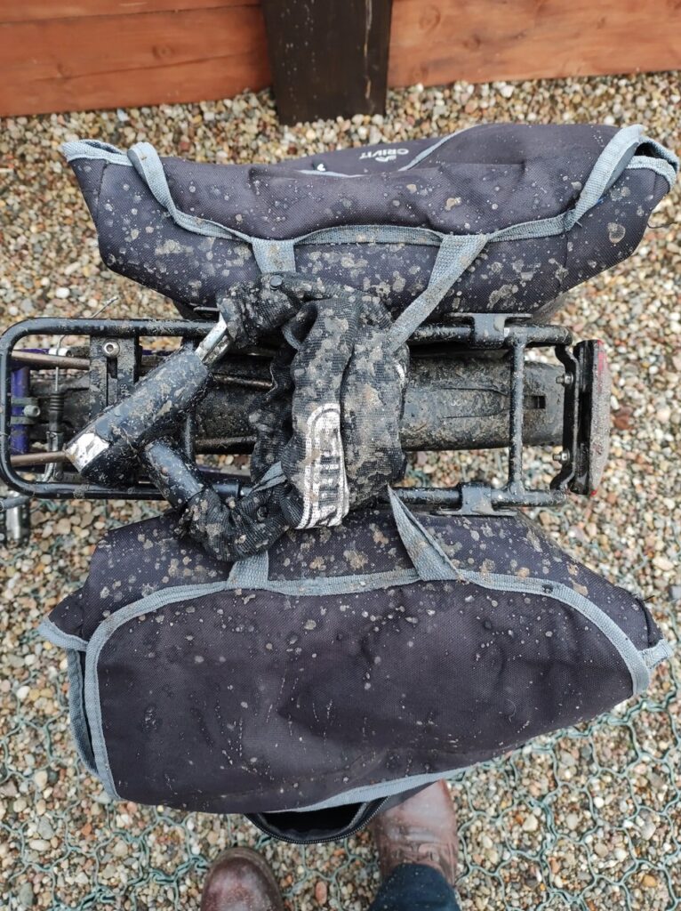 Widziane z góry czarne sakwy zamontowane do bagażnika roweru i spięte razem z nim solidnym łańcuchem firmy Abus. Błotnik pod bagażnikiem, wierzch sakw i łańcuch są pobrudzone błotem.