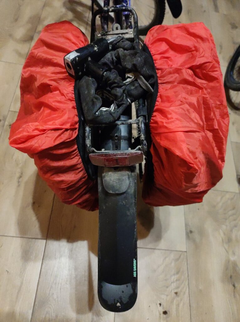 Sakwy na bagażniku widzianego od tyłu roweru, tym razem w czerwonych przeciwdeszczowych pokrowcach. Błotnik ledwo wystający spod bagażnika został przedłużony przez długi kawałek czarnego plastiku.