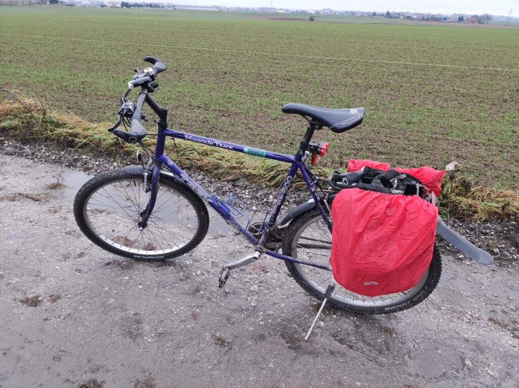 Ciemnoniebieski rower trekingowy Trek stoi na błotnistej polnej drodze. Na bagażniku ma sakwy w czerwonych pokrowcach, z tyłu wystaje mocno przedłużony błotnik.