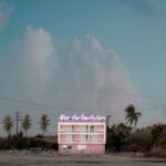 Nieduży, dwupiętrowy, różowo-biały budynek z różowym neonem „After the Revolution” na tle palm i krzewów.