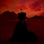 Na zdjęcie czerwonej pustyni z ciemniejszymi sylwetkami wzgórz w tle nałożona jest Nubya Garcua w długiej czarnej sukni, trzymająca w rękach sznury pereł. Jest oświetlona tak, że większość skrywa się w ciemności, a tylko oczy są w świetle.