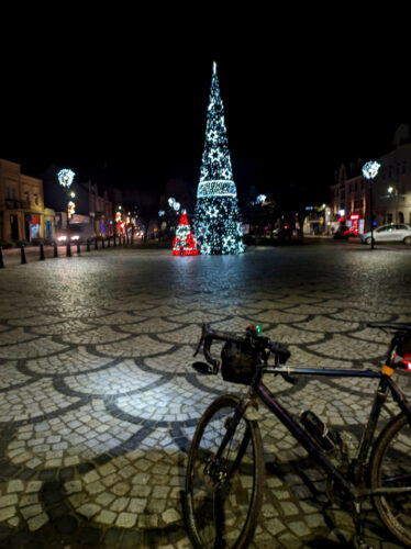 Na pierwszym planie stoi ciemnoniebieski rower szutrowy, dalej brukowana płyta rynku, na której stoi świecąca choinka, a po jej lewej stronie świecący kolorowy mikołaj.