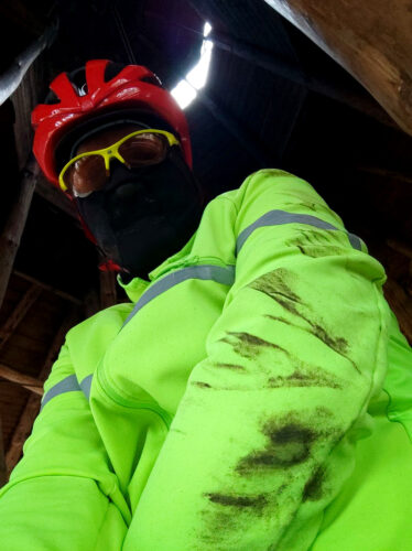 Selfie zrobione z lewej strony i z poziomu pasa, żeby było widać pobrudzony przy wywróceniu lewy rękaw jaskrawej kurtki rowerowej. Mam na sobie kominiarkę, okulary rowerowe i czerwony kas.