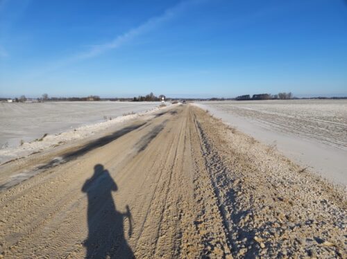 Słoneczny dzień. Pod intensywnie błękitnym niebem znajduje się szosa przez pola zasłonięta jasnoszarą breją pośniegową, częściowo zmarznięta w koleiny. W kilku miejscach szosy śnieg jest wyjeżdżony i zastąpiony przez fragmenty lodu.