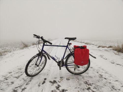 Ciemnogranatowy rower Trek z dużymi sakwami na bagażniku osłoniętymi czerwonymi pokrowcami stoi w poprzek zaśnieżonej polnej drogi w bardzo mglisty dzień.