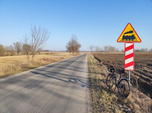 Kolejne zdjęcie roweru, tym razem opartego o znak ostrzegający o niestrzeżonym przejeździe kolejowym, widocznym w głębi zdjęcia.
