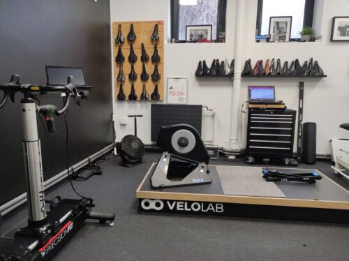 Sala bike fittingu. Po lewej widać fragment urządzenia pozwalającego na odtworzenie roweru o dowolnej geometrii z możliwością przesunięcia oraz podwyższenia kierownicy, siodła itp. Na wprost na podeście z logo VeloLab znajduje się pusty trenażer Tacx. Na ścianie za nim sporo rowerowych siodełek. Poza tym laptop, wentylator, szafka (najprawdopodobniej) na części, poziomica i oprawione czarno-białe pamiątkowe zdjęcia.