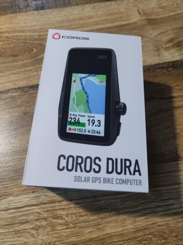 Na drewnianym stole leży jasnoszare pudełko z komputerkiem rowerowym GPS Dura z firmy Coros. Na środku pudełka znajduje się zdjęcie urządzenia, nad nim logo firmy, a na spodzie nazwa sprzętu z dopiskiem „Solar GPS bike computer”.