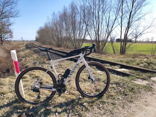 Słoneczny dzień. Jasnokremowy rower szutrowy Canyon Grizl stoi oparty tylnym kołem o przydrożny słupek tuż przy torach kolejowych skręcających w oddali w prawo. Pomiędzy krzakami widać zielone pola i błękitne niebo.