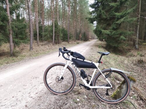 Ten sam rower szutrowy, tym razem wyposażony jest już w torby. Jedna mała znajduje się na ramie, a większa jest wpasowana w ramę. Rower toi oparty o omszały, betonowy słupek przy szutrowej drodze przez las.