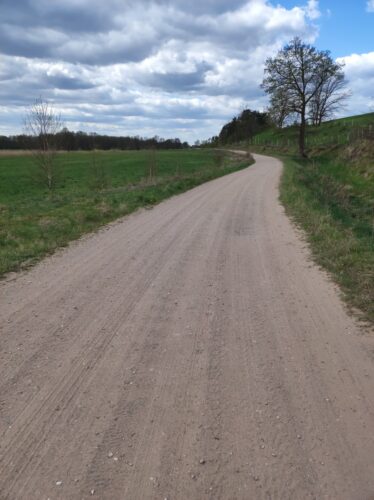 Skręcająca w lewo i prowadząca w stronę widocznego w oddali lasu droga szutrowa pod pochmurnym niebem.