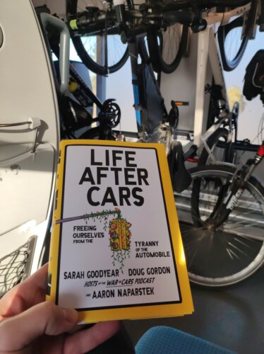 Moja ręką trzymająca książkę "Life After Cars. Freeing Ourselves from the Tyrrany of the Automobile” Sarah Goodyear, Douga Gordona i Aarona Naparstka, a w tle rowery wiszące i stojące w wagonie.