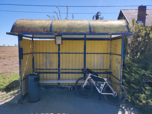 O ławkę na żółto-niebieskim przystanku autobusowym oparty jest jasno-kremowy, prawie biały rower szutrowy Canyon Grizl.