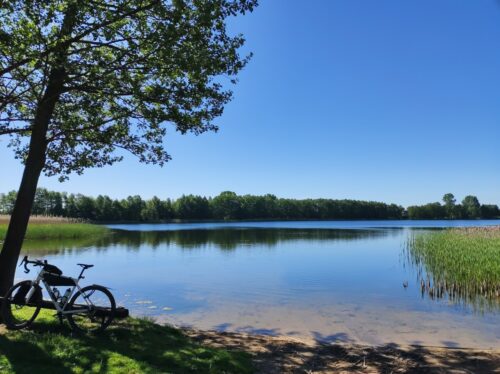 Jezioro widziane z niedużej plaży. Po lewej, oparty o ławkę przy drzewie, stoi mój rower.