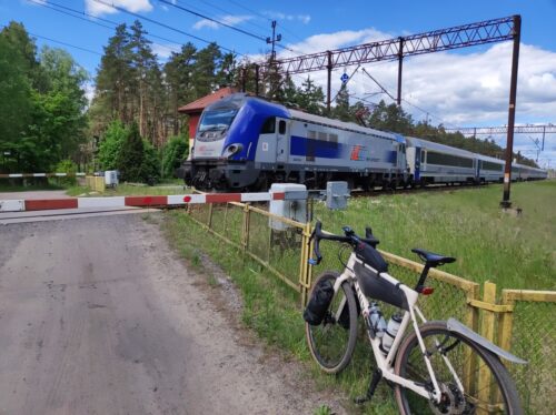 Pociąg Intercity wjeżdża na zamknięty przejazd, przed który stoi mój rower, oparty o żółty płot.