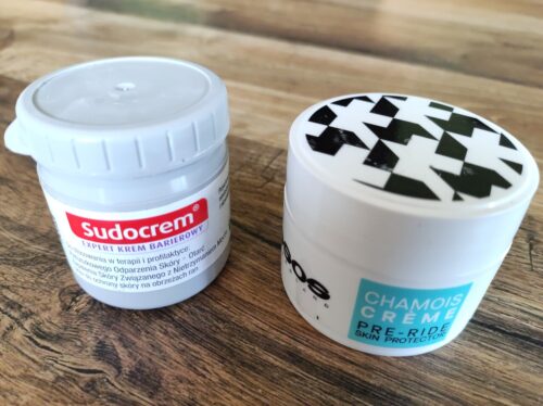 Dwa plastikowe słoiczki z kremem. Po lewej szary z Sudocremem, po prawej biały z Assos Chamois Creme.