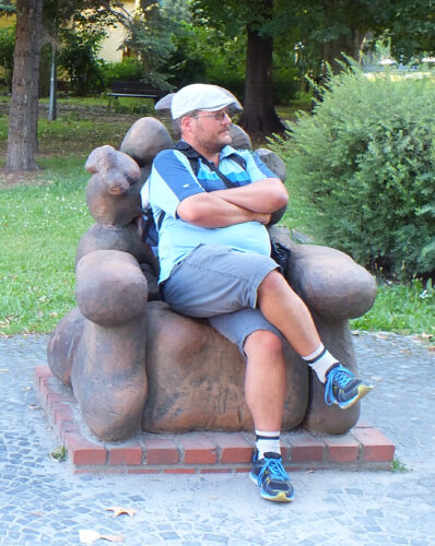 Siedzę na rzeźbie – fotelu z kamienia. Mam na sobie spodenki do kolan, niebieską koszulkę polo i kaszkiet. Koszulka jest wypchana moim brzuchem, który układa się na kolanach.