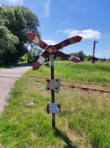 Zniszczony znak ostrzegający przed przejazdem kolejowym. Biało-czerwony krzyż świętego Andrzeja jest zardzewiały i ma pogięte ramiona, znajdujący się poniżej znak STOP jest niemalże nierozpoznawalny, bo zostało z niego jedynie TO. Na samym dole jest tabliczka z napisem „STRZE SIĘ OCIĄGU”.