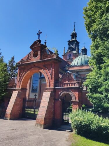 Duży kościół z czerwonej cegły widziany zza bramy. Kopuła nad prawą częścią oraz zwieńczenie wierzy są z oksydowanej na zielono miedzi.