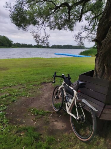 Mój jasnokremowy rower szutrowy Canyon Grizl opiera się o drewnianą ławkę otaczającą drzewo rosnące na brzegu jeziora. W tle jezioro i niebieski pływający pomost.